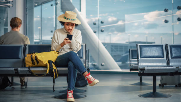 terminal de l’aéroport: une femme noire attend son vol, utilise un smartphone, reçoit des nouvelles scandaleusement mauvaises, manque le vol. personne bouleversée, triste et déçue assise dans une salle d’embarquement d’un hub aérien. - salle dembarquement photos et images de collection