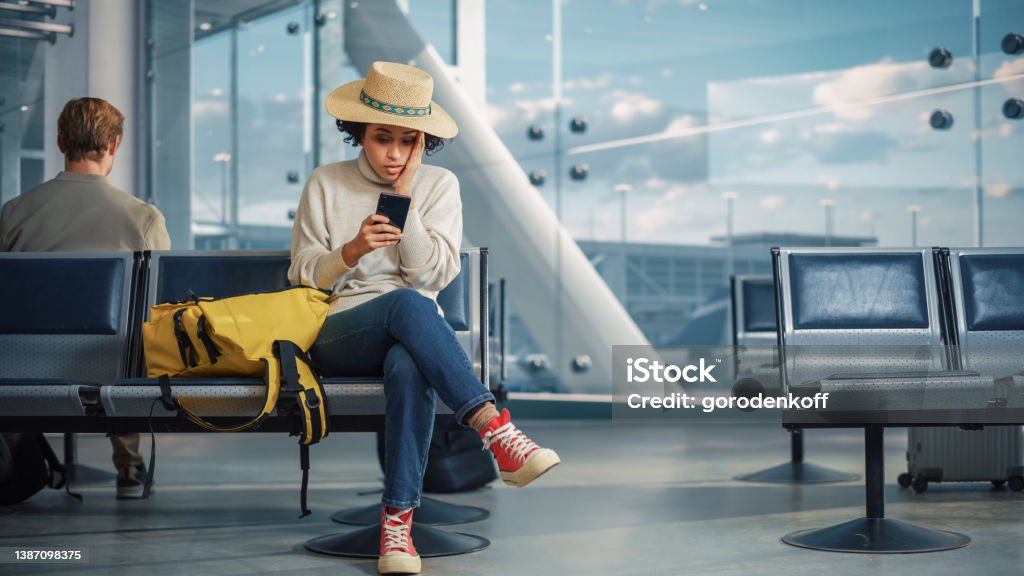 Airport Terminal: Black Woman Waits for Flight, Uses Smartphone, Receives Shockingly Bad News, Misses Flight. Upset, Sad, and Dissappointed Person Sitting in a Boarding Lounge of Airline Hub. Commercial Airplane Stock Photo