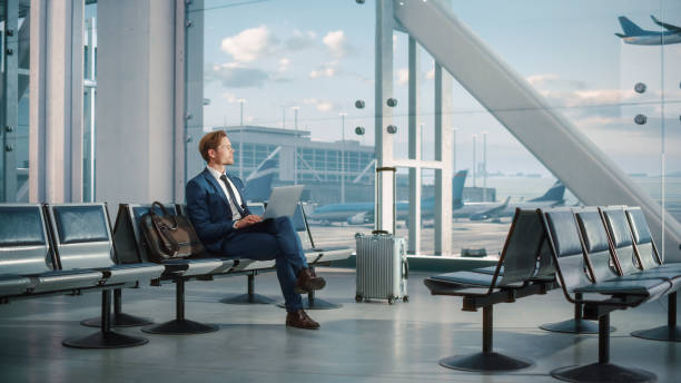 istock Airport Terminal: Businessman Uses Laptop Computer, Waiting for His Flight. Traveling Entrepreneur Remote Work Online Sitting in Boarding Lounge of Airline Hub with Airplanes Arriving, Departing. Wide 1387099072