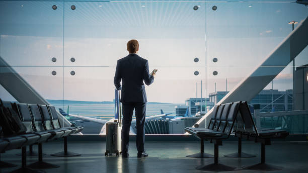 istock Airport Terminal: Businessman with Rolling Suitcase Walks, Uses Smartphone App for e-Business. Back View Silhouette of Traveling Man Waits for Flight in Boarding Lounge of Airline Hub with Airplanes 1387099112