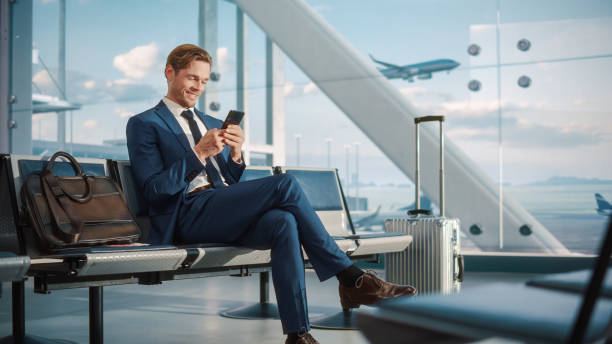 istock Airport Terminal Flight Wait: Smiling Businessman Uses Smartphone for e-Business, Browsing Internet with an App. Traveling Entrepreneur Work Online on Mobile Phone in Boarding Lounge of Airline Hub 1387099203