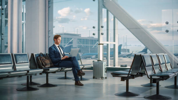 istock Modern Airport Terminal: Handsome Businessman Working on Laptop Computer While Waiting for His Flight. Man Sitting in a Boarding Lounge of International Airline Hub with Airplanes Departing, Arriving 1387099302