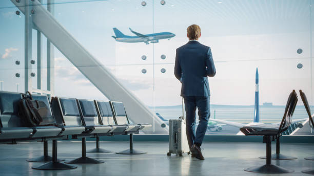 istock Airport Terminal: Businessman with Rolling Suitcase Walks, Uses Smartphone App for Internet e-Business. Traveling Entrepreneur Work Online while Waiting for a Flight in Boarding Lounge of Airline Hub 1387099341