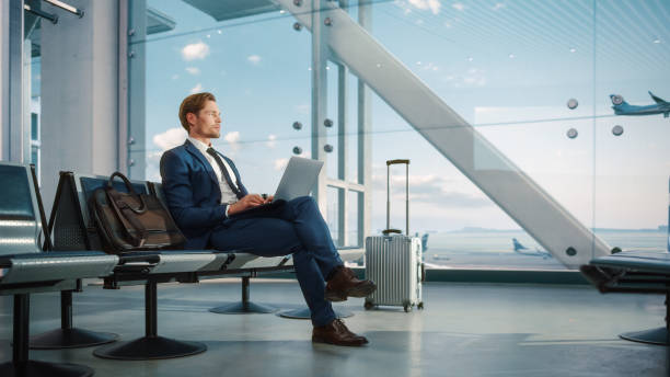 istock Airport Terminal: Businessman Uses Laptop Computer, Waiting for Flight. Traveling Man Relax after Work, Looking forward to Vacation. Sitting in a Boarding Lounge of Airline Hub with Airplanes 1387099352