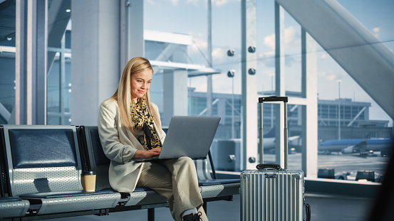 Airport Terminal: Smiling Woman Waits for Flight, Uses Laptop, Browse Internet, does e-Business, Online Shopping. Traveling Female Remote Work Online on Computer in a Boarding Lounge of Airline Hub