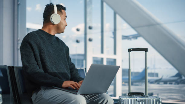 Airport Terminal: Businessman Uses Laptop Computer, Listents to Musics or Podcast in Headphone, Waiting for Flight. Traveling Digital Entrepreneur Remote Work Online in Boarding Lounge of Airline Hub terminal del aeropuerto: hombre de negocios usa computadora portátil, escucha músicas o podcasts en auriculares, esperando el vuelo. emprendedor digital viajero trabajo remoto en línea en la sala de embarque del centro de operaciones de la aerolínea