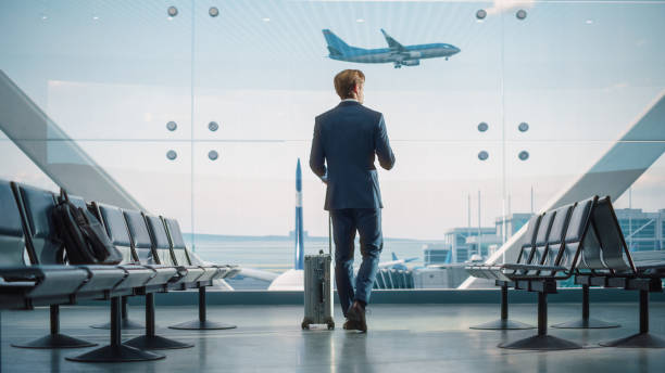 istock Airport Terminal: Businessman with Rolling Suitcase Walks, Uses Smartphone App for e-Business. Back View of Traveling Man Waiting for a Flight in Boarding Lounge of Airline Hub with Airplanes Arriving 1387099519