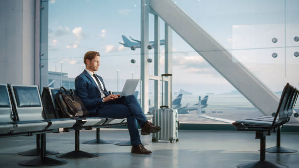 istock Modern Airport Terminal: Handsome Businessman Working on Laptop Computer While Waiting for His Flight. Man Sitting in a Boarding Lounge of Big Airline Hub with Airplanes Departing and Arriving 1387099547