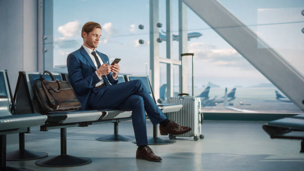 istock Airport Terminal: Businessman Uses Smartphone, Waiting for a Flight, Doing e-Business, Browsing the Internet with an App. Traveling Entrepreneur Remote Work Online in Boarding Lounge of Airline Hub 1387099616
