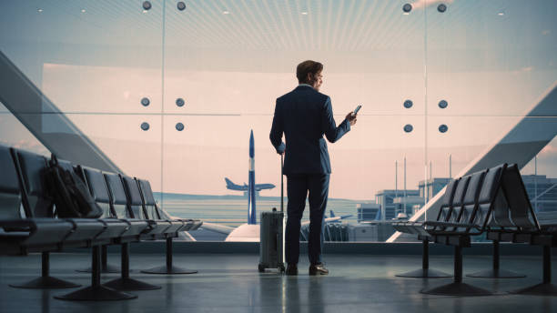 istock Airport Terminal: Businessman with Rolling Suitcase Walks, Uses Smartphone App for e-Business. Back View Silhouette of Traveling Man Waits for Flight in Boarding Lounge of Airline Hub with Airplanes 1387099755