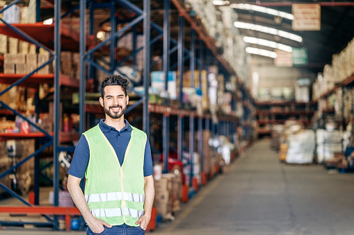 Portrait of a happy young warehouse worker wearing reflective vest. Smiling male manual worker standing with his hands in pockets in large distribution warehouse.
