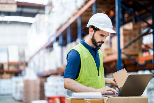 istock Man holding a box and working in laptop in warehouse 1387105344