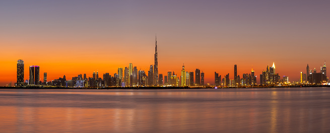 Panorama of Dubai Business Bay skyline at night after sunset with colorful illuminated buildings and calm Dubai Creek water.