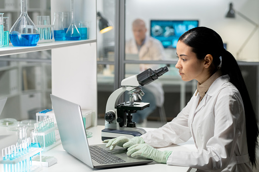 Young contemporary female chemist in whitecoat and gloves typing on laptop keyboard while looking at screen in laboratory