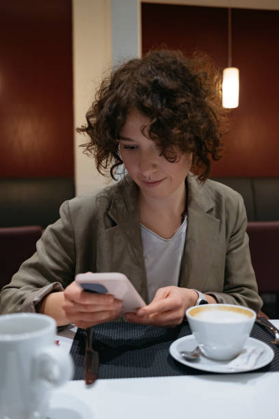 une femme d’affaires heureuse assise dans un restaurant, déjeunant et buvant un café après une longue journée de travail; vérifier sur son smartphone s’il y a un point d’accès wi-fi ouvert - facilité daccès photos photos et images de collection