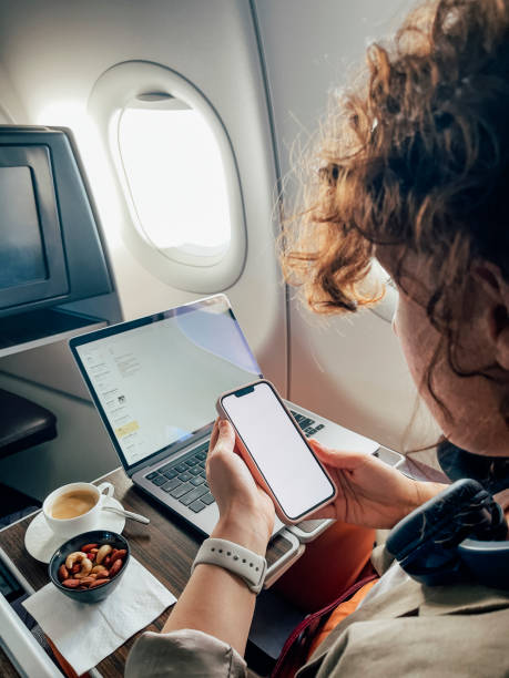An Unrecognizable Caucasian Woman Sitting In The Airplane And Using Her Smartphone While Her Laptop Is On In The Background