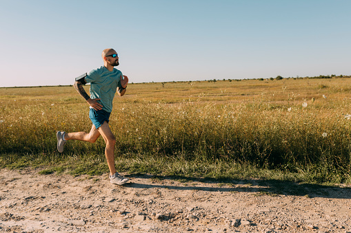 A fit white sportsman wearing sunglasses running in the field on a sunny day.