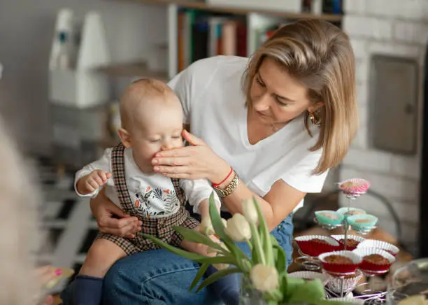 Beautiful blonde child choked. Young blonde mom wipes her baby dirty mouth. The child got dirty while eating. Casual clothed family having lunch together, eating cupcakes at home indoors.