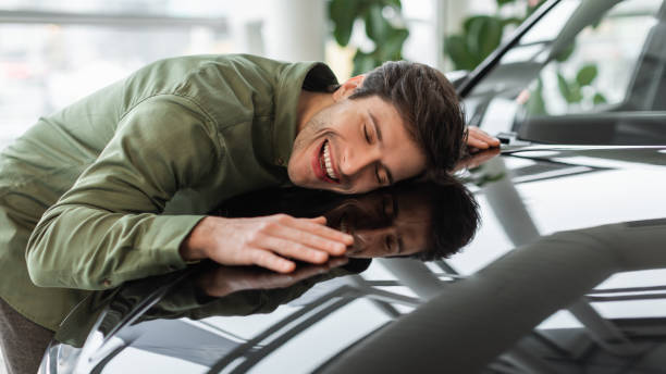 Happy young man hugging hood of his new car, dreaming about buying vehicle at auto showroom, panorama stock photo