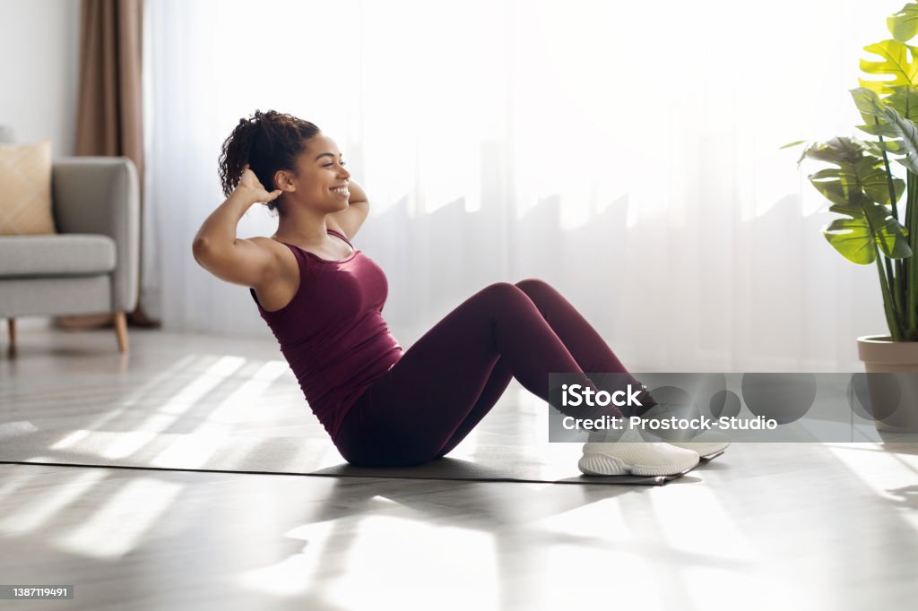 Cheerful african american woman exercising on fitness mat at home Cheerful millennial african american woman in sportswear exercising on fitness mat at living room alone, doing crunches, working on abs, looking at copy space and smiling, side view. Sport at home Sit-ups Stock Photo