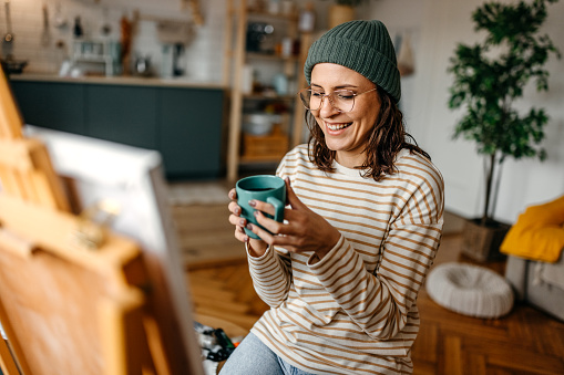 Smiling young woman wearing knit hat and eyeglasses holding coffee cup while relaxing at home