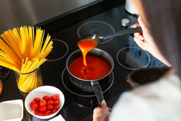 Photo of High angle, over the shoulder view of a woman stirring boiling soup from Saucepanwith tomato