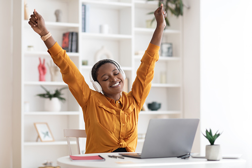 Happy stylish short-haired young african american woman employee having break while working on laptop at office, sitting at workdesk with closed eyes, using wireless headphones, raising hands up