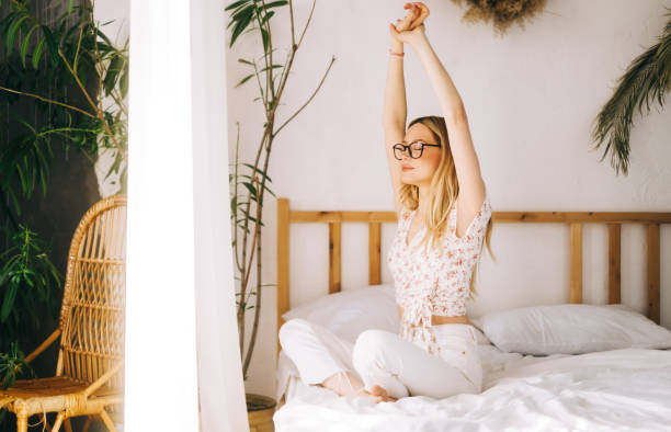 atrakcyjna kaukaska młoda kobieta w okularach medytuje siedząc na łóżku i ciesząc się słonecznym porankiem. - yoga young adult cheerful happiness zdjęcia i obrazy z banku zdjęć