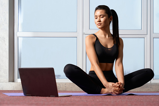 Front view of brunette girl sitting on legs, relaxing indoors. Young sporty female practicing yoga, having break, looking at laptop, holding by crossed fingers. Concept of yoga.