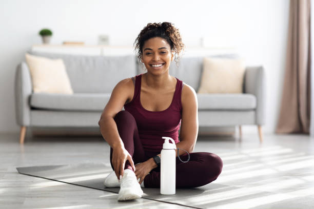 femme noire sportive positive assise sur un tapis de fitness et souriante - vêtement de sport photos et images de collection