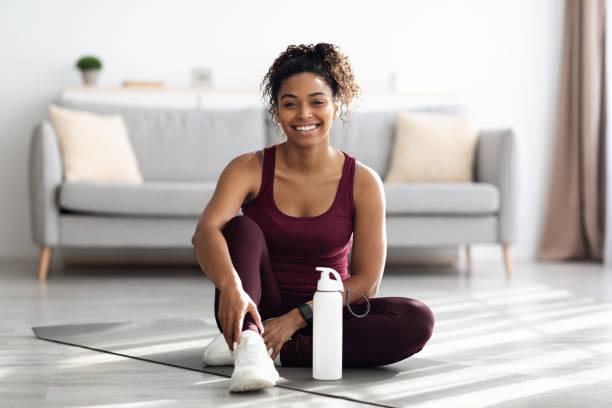 donna nera sportiva positiva seduta sul tappetino fitness e sorridente - sports clothing foto e immagini stock
