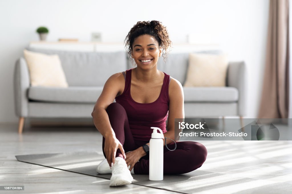 Positive sporty black lady sitting on fitness mat and smiling Positive sporty millennial black lady in beautiful sportswear and white sneakers sitting on fitness mat with bottle of water and smiling, having break while exercising at home, copy space Women Stock Photo