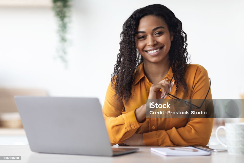 Pretty african american woman freelancer using laptop at home Pretty long-haired millennial african american woman freelancer using laptop at home, black lady sitting at table at kitchen, holding eyeglasses and smiling at camera, copy space. Remote job concept Women Stock Photo