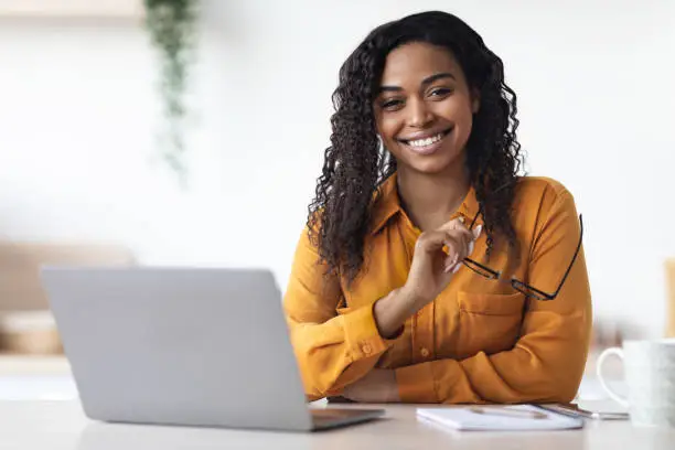 Pretty long-haired millennial african american woman freelancer using laptop at home, black lady sitting at table at kitchen, holding eyeglasses and smiling at camera, copy space. Remote job concept