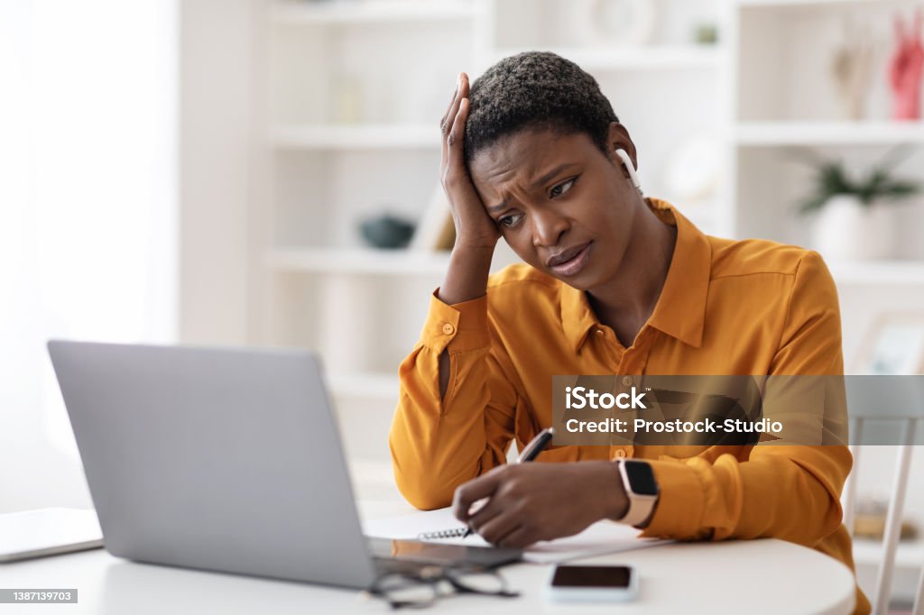 Bored black lady attending webinar, sitting in front of laptop Bored stylish short-haired young black lady attending webinar, sitting in front of laptop, using wireless earbuds, taking notes, looking at computer screen and touching her head, copy space Boredom Stock Photo