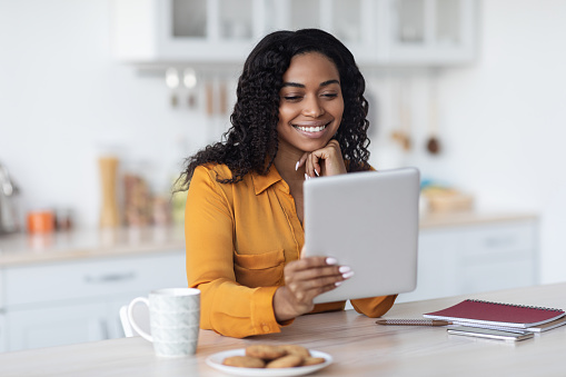 Happy pretty millennial black woman in casual outfit using modern digital tablet, having coffe break, surfing on social media, having video call, kitchen interior, copy space. Gadget addiction concept