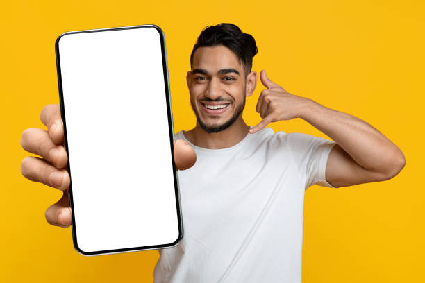 Happy arab guy showing cell phone with blank screen Happy attractive bearded arab guy in white t-shirt showing modern cell phone with white blank screen and call me gesture, smiling at camera, yellow studio background, mockup free betting advice stock pictures, royalty-free photos & images