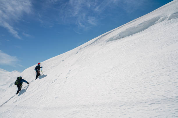 Mature Caucasian male mountaineers are ascending a snowy slope with determination under a clear blue sky. Their colorful gear contrasts with the pristine snow, highlighting their adventurous spirit and teamwork in this exhilarating mountain environment.