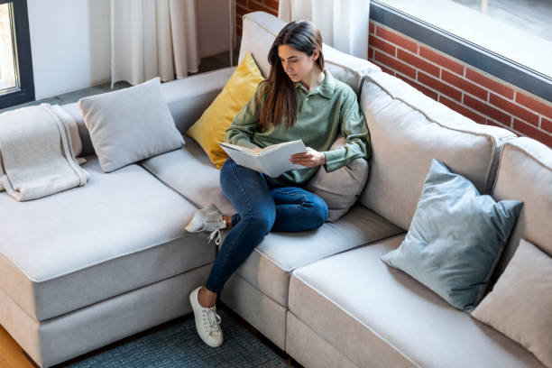 istock Pretty young woman reading a book while sitting on sofa at home. 1387145946