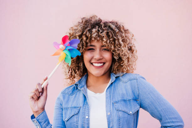 beautiful happy hispanic woman with afro hair holding colorful pinwheel. pink background,wind energy stock photo
