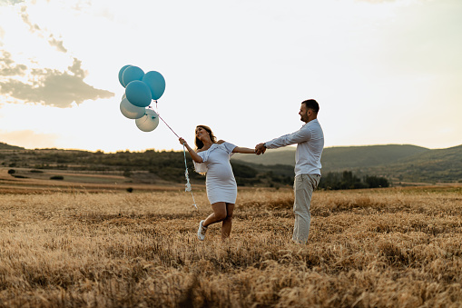 Happy pregnant woman and a man revealing baby gender with blue balloons on a sunny summer day in nature
