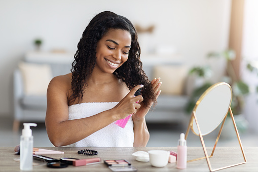 Pretty african american millennial lady taking care about her curly hair, sitting in front of mirror covered in towel, brushing hair, using product against split ends and smiling, living room interior