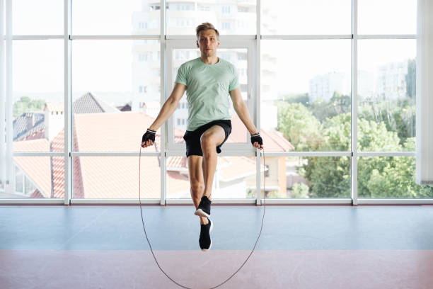 Handsome young man jumping over skipping rope working out Athletic young man training with skipping rope. Cardio workout with minimal equipment in a big space with a lot of daylight and glass wall skipping stock pictures, royalty-free photos & images
