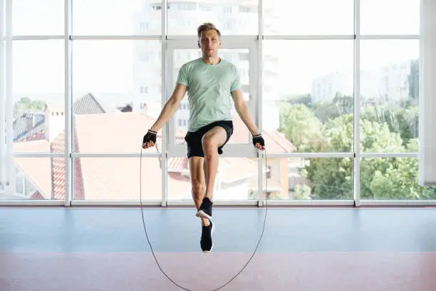 Athletic young man training with skipping rope. Cardio workout with minimal equipment in a big space with a lot of daylight and glass wall
