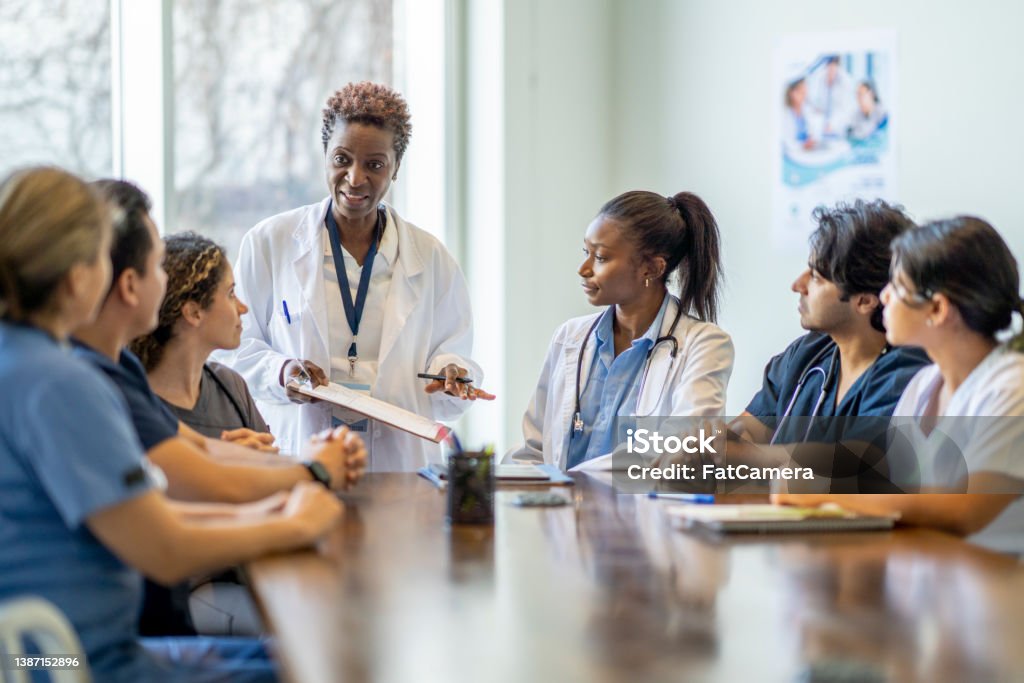 Female Doctor Teaching Nursing Students A small group of diverse nursing students sit around a boardroom table as they listen attentively to their teacher and lead doctor.  They are each dressed in medical scrubs and sitting with papers out in front of them.  The doctor is holding out a clipboard with a document on it as she reviews it with the group. Healthcare And Medicine Stock Photo