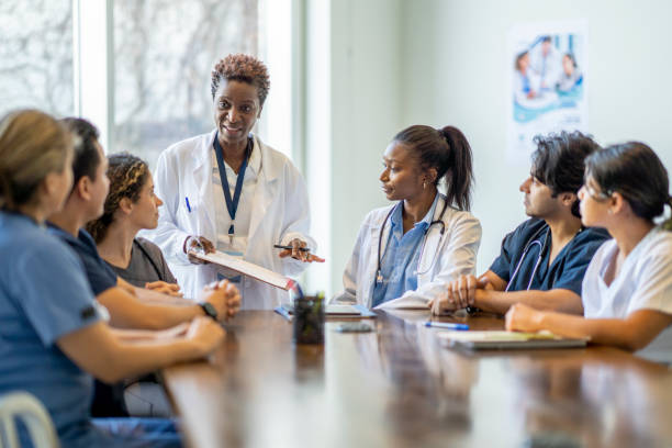 Female Doctor Teaching Nursing Students A small group of diverse nursing students sit around a boardroom table as they listen attentively to their teacher and lead doctor.  They are each dressed in medical scrubs and sitting with papers out in front of them.  The doctor is holding out a clipboard with a document on it as she reviews it with the group. medical education stock pictures, royalty-free photos & images