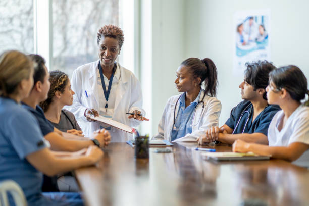 Female Doctor Teaching Nursing Students A small group of diverse nursing students sit around a boardroom table as they listen attentively to their teacher and lead doctor.  They are each dressed in medical scrubs and sitting with papers out in front of them.  The doctor is holding out a clipboard with a document on it as she reviews it with the group. public health stock pictures, royalty-free photos & images