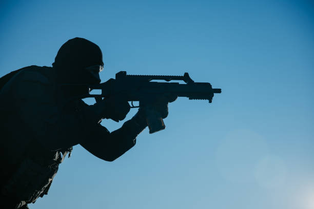 Male soldier with a rifle wearing balaclava, he is aiming under a blue sky.