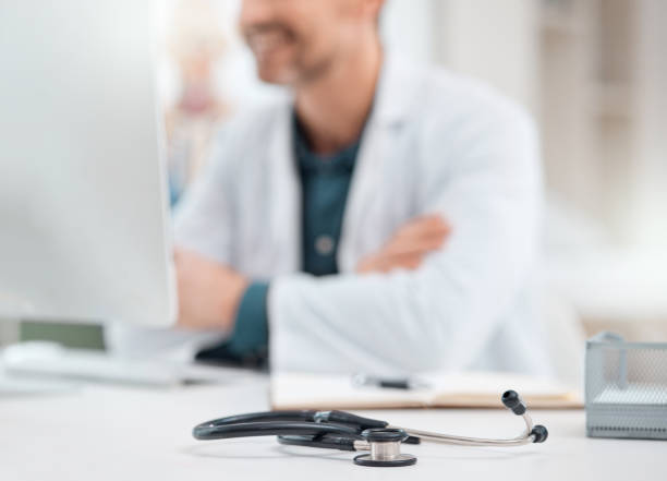 istock Closeup shot of a stethoscope on a table with a doctor working on a computer in the background in a medical office 1387161484