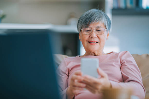 donna anziana cinese asiatica che utilizza il messaggio di testo di lettura dello smartphone nel soggiorno del suo appartamento in città - navigare in internet foto e immagini stock